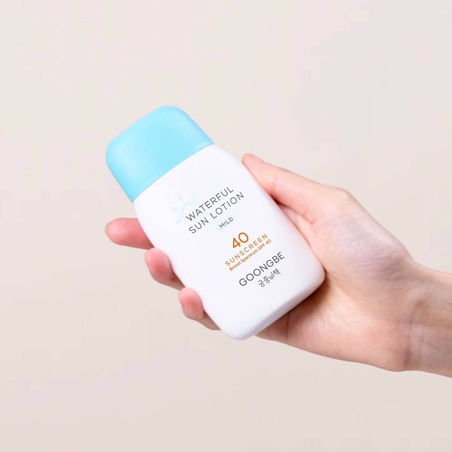 Hand holding a bottle of 'GOONGBE' sun lotion with a light blue cap on a beige background