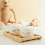 Two containers of GOONGBE skincare product on a wooden tray with a soft, blurred background.